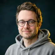 Mies silmälasit päässä ja harmaa huppari yllään istuu tietokoneen ääressä